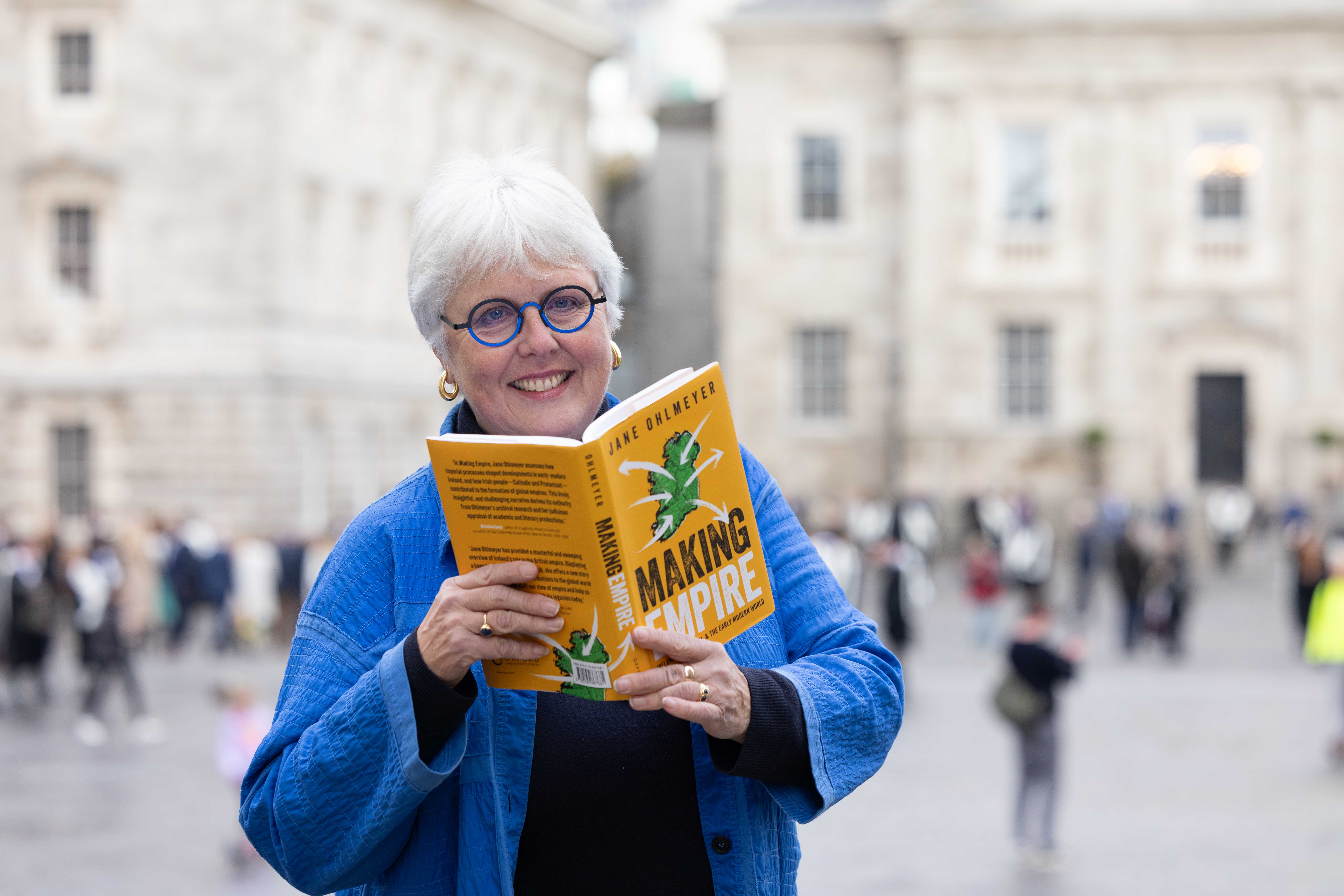 Portrait photo of Jane Ohlmeyer holding her book, 'Making Empire,' with classical buildings and people in the background