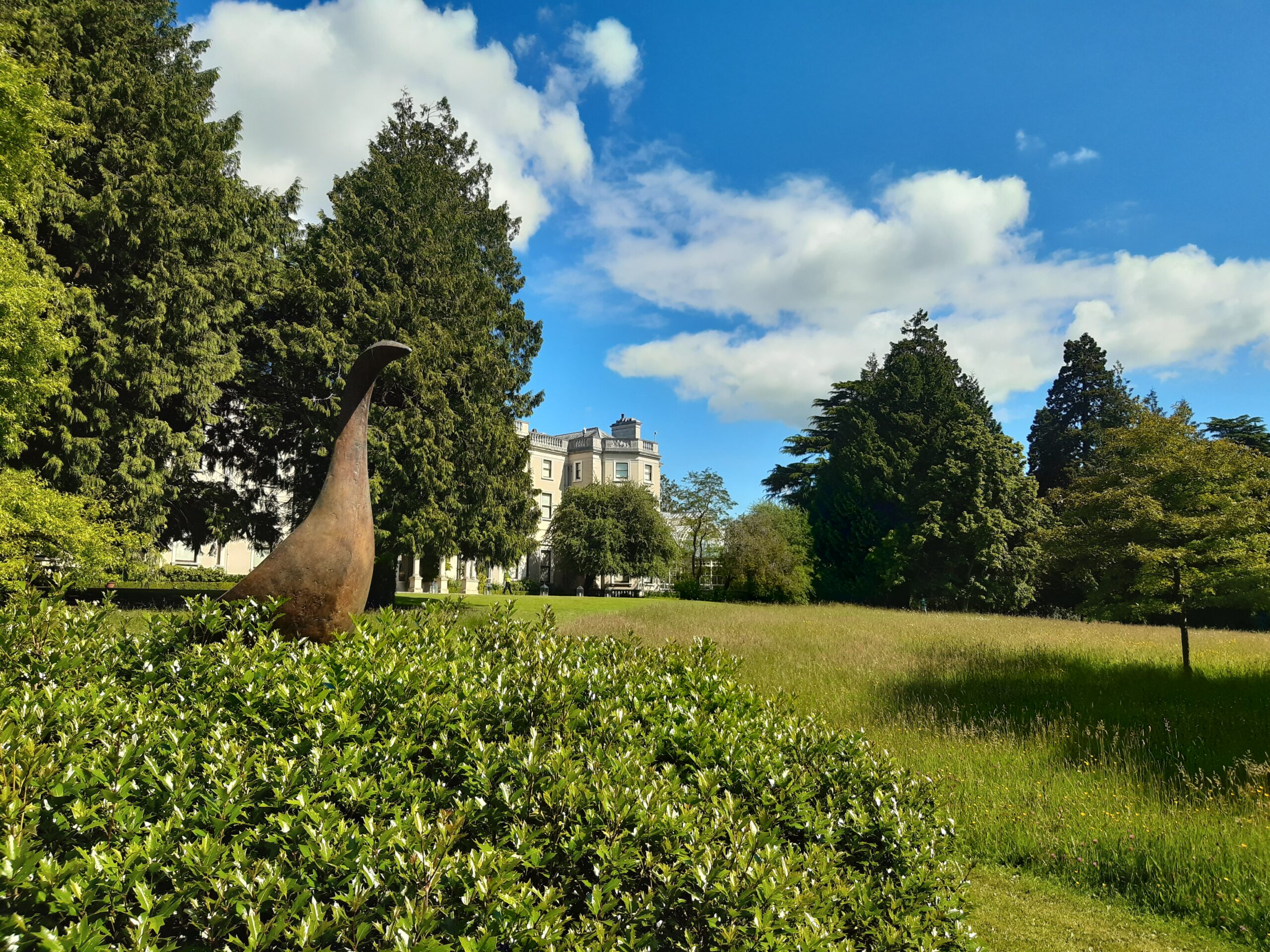 Sculpture of bronze bird on lawn in front of Farmleigh House, house partially obscured by trees.