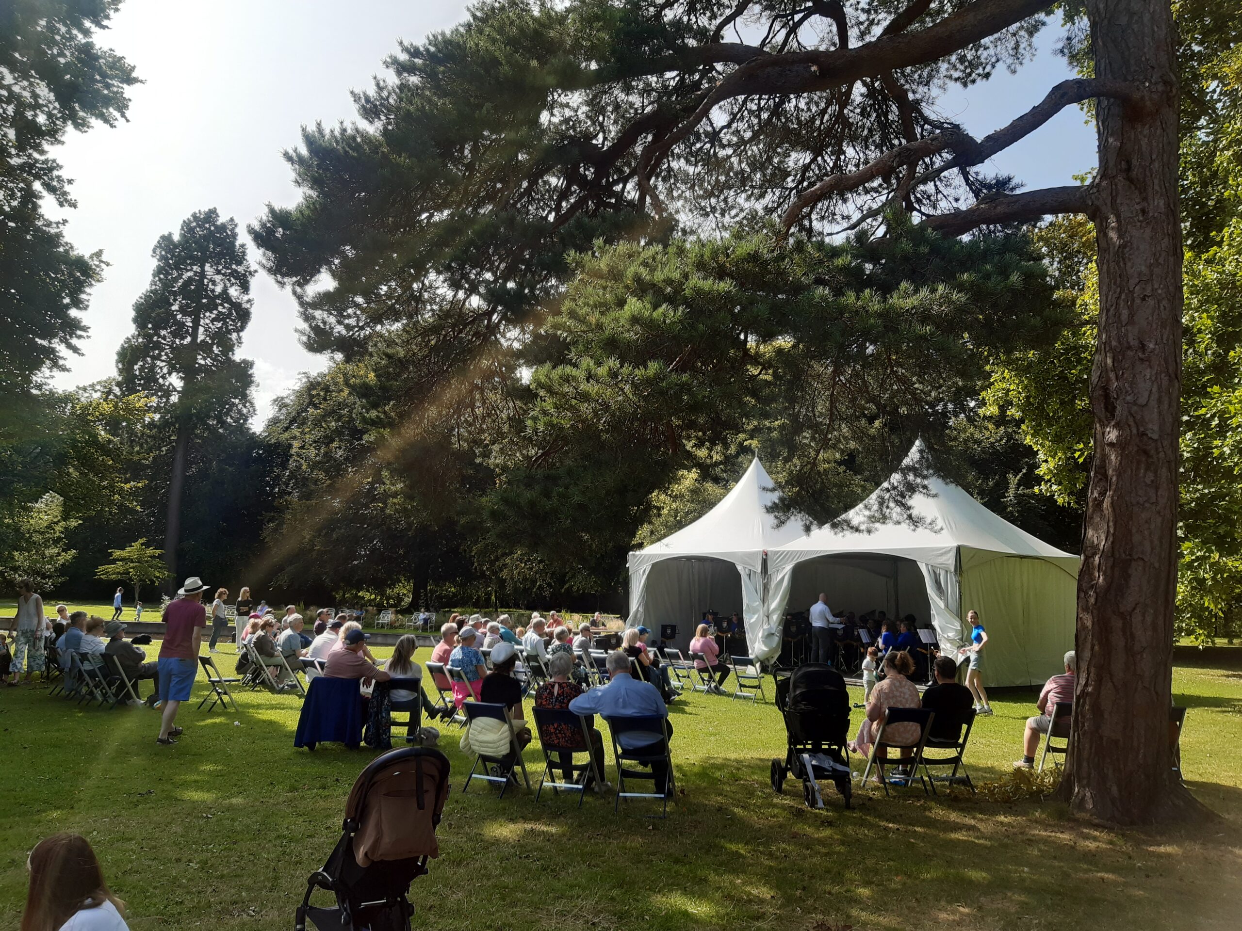 Brass band in white tent entertaining visitors on lawn surrounded by mature trees.